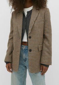 Blazer à motif pied-de-poule marron et beige avec un col à revers cranté, deux boutons noirs, et des poches plaquées, porté par-dessus un cardigan gris et un jean.