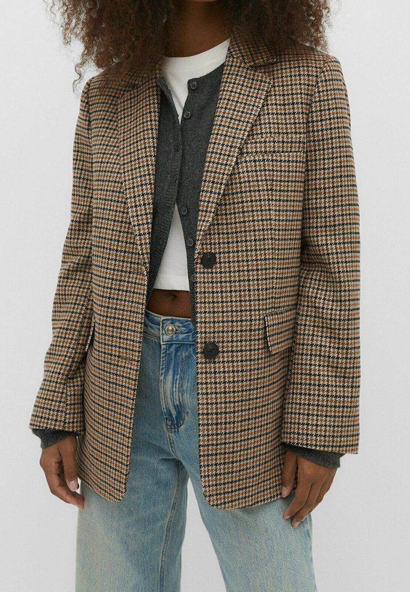 Blazer à motif pied-de-poule marron et beige avec un col à revers cranté, deux boutons noirs, et des poches plaquées, porté par-dessus un cardigan gris et un jean.