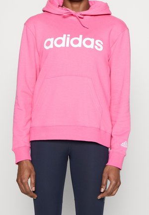 Person trägt einen knallpinken Adidas-Hoodie mit weißem Logo und dunkelblauen Leggings und steht vor einem schlichten hellen Hintergrund.
