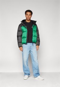 Chaqueta acolchada con un frente verde y mangas negras, que incluye capucha, cierre de cremallera y textura acolchada, combinada con jeans azul claro y zapatillas blancas.