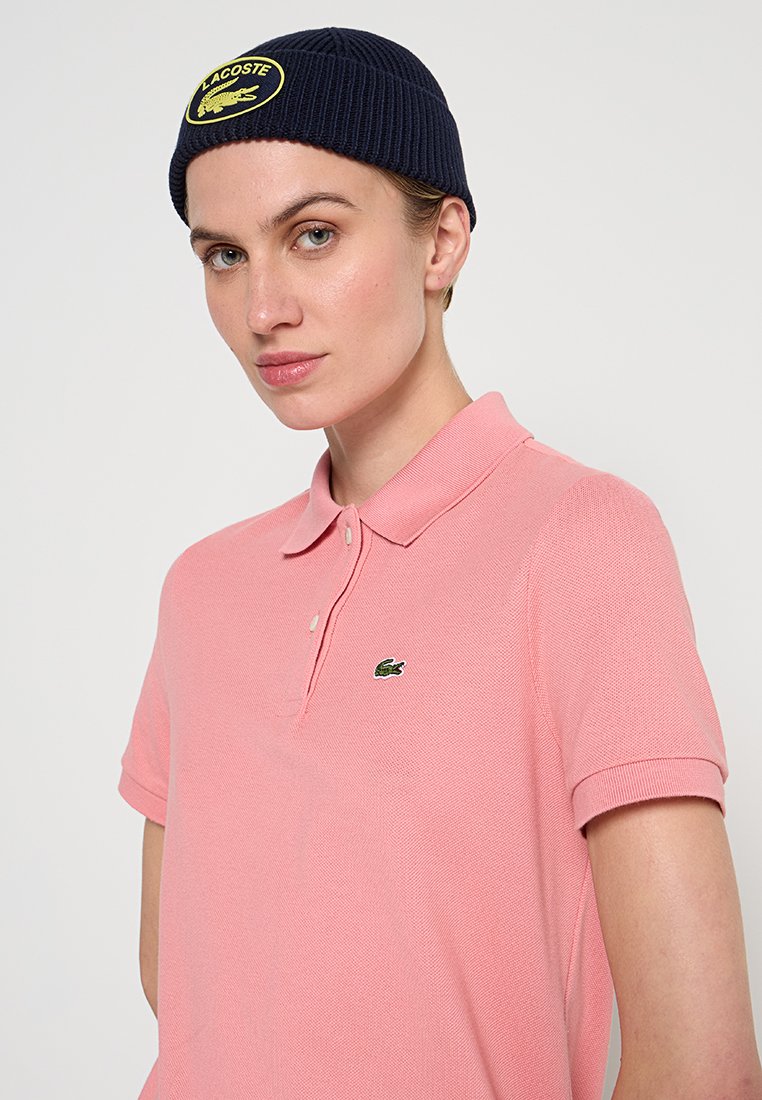 Polo en coton rose clair, avec un col classique et une patte de boutonnage à deux boutons, offrant un logo crocodile brodé vert sur la poitrine.