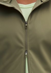 Giacca a zip verde oliva con una texture di tessuto morbido, chiusura a doppio zip e uno strato verde chiaro sotto.