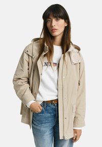 Beige letvægtsjakke med hætte, sølv hardware og frontlommer, båret over en hvid sweatshirt og blå jeans.