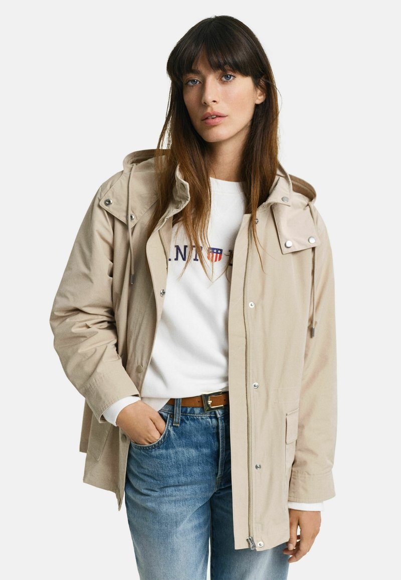 Beige letvægtsjakke med hætte, sølv hardware og frontlommer, båret over en hvid sweatshirt og blå jeans.