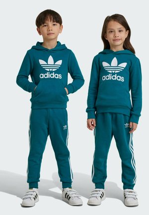 Sudadera y pantalones deportivos Adidas en teal con rayas blancas; presenta el icónico logo en el frente. Dos niños posan con el atuendo, mostrando un diseño deportivo.