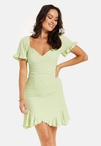 Green polka dot dress with short ruffled sleeves, V-neckline, fitted bodice, and a flared hem with ruffle detail, made of lightweight fabric.