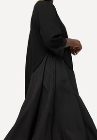 Černý dvoudílný oblek s krčkovým topem s tříčtvrtečními rukávy a dlouhou, splývavou sukní. Texturované akcenty na rukávech a hladká tkanina.