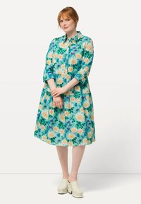 Vestito button-up floreale in turchese con fiori crema e gialli, design a strati, maniche a tre quarti, abbinato a zoccoli bianchi.