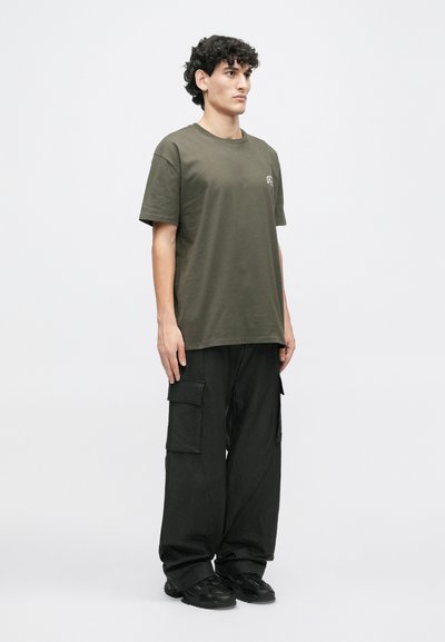 Olivnozeleni bombažni majica s krojem okroglega vratu, v kombinaciji z ohlapnimi črnimi cargo hlačami, ki imajo več žepov in teksturiran zaključek.