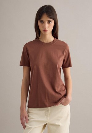 Donna con capelli castani lisci, indossa una maglietta marrone a maniche corte e pantaloni beige, in piedi contro uno sfondo neutro semplice.