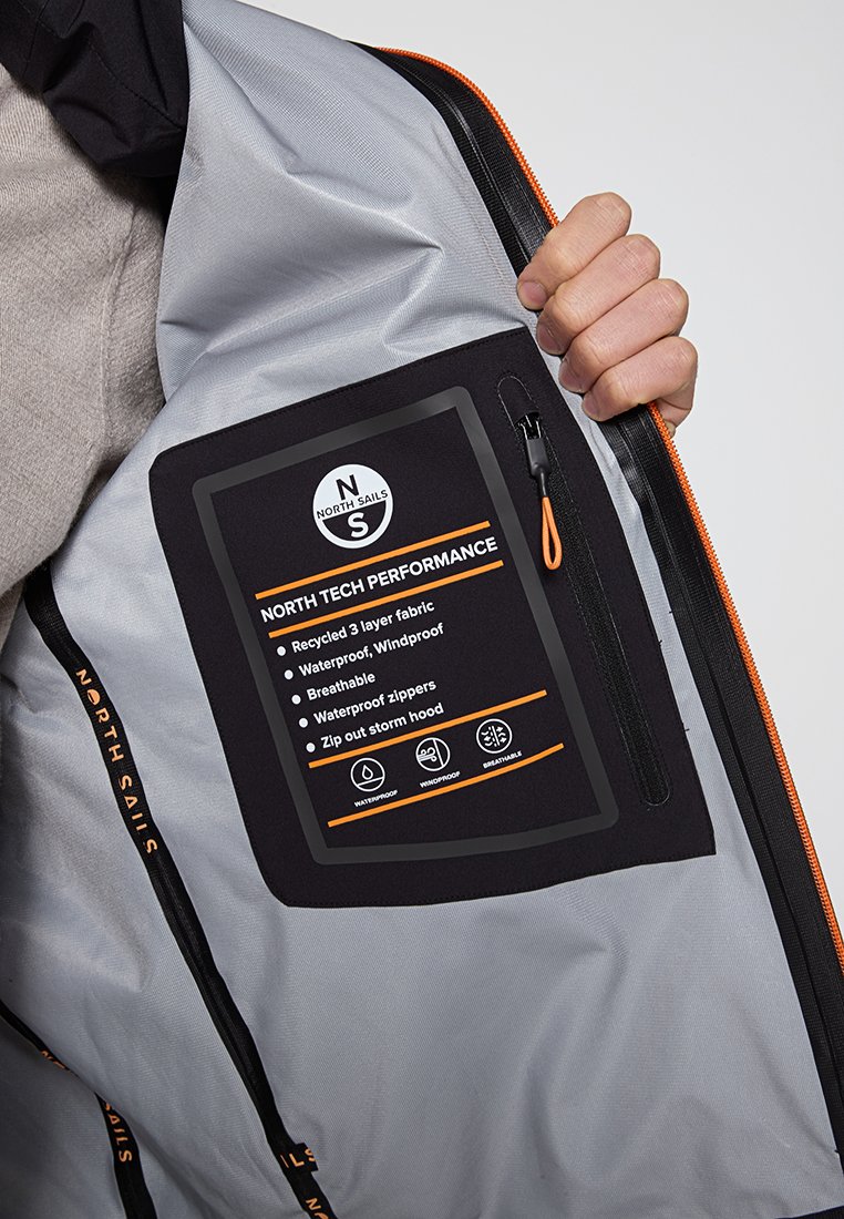 Mano che tiene aperto l'interno di una giacca grigia mostrando un'etichetta che dettaglia il tessuto impermeabile, antivento e traspirante North Sails con cappuccio ant tempesta removibile.