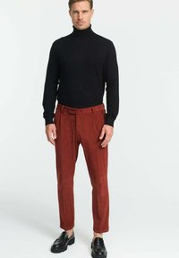 Schwarzer Rollkragenpullover kombiniert mit rostfarbenen Kordhosen. Die Hosen verfügen über einen Knopfverschluss und vertikale Rillen. Schwarze Mokkasins runden das Outfit ab.