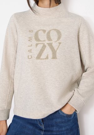 Sweat-shirt beige avec un texte texturé « CALM & COZY » porté par une personne tenant la main dans la poche, associé à un jean bleu.