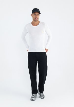 Vit långärmad skjorta med rund halsringning och liten gul emblem, i kombination med svarta byxor och grå sportskor; person som bär en svart mössa.