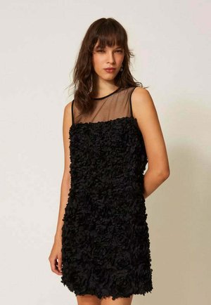 Donna con un abito nero senza maniche in tessuto floreale strutturato e scollo trasparente, in piedi davanti a uno sfondo chiaro e uniforme.