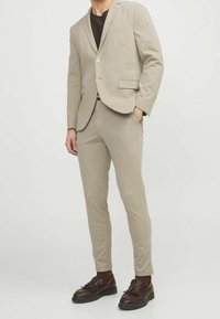 Béžový oblek vyrobený z měkké tkaniny, s blejzrem na jedno knoflíkové zapínání a slim-fit kalhotami. Outfit doplňují hn ědé boty.