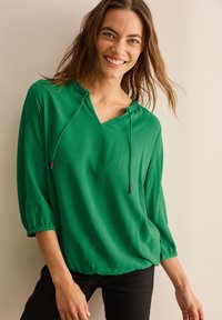 Blusa verde leggera con scollo a V e caratteristica con cordoncino. Ha maniche a tre quarti e una vestibilità rilassata, abbinata a pantaloni neri.