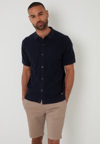 Pull à manches courtes bleu marine avec un design en câble texturé, devant boutonné et col plat, associé à un short beige clair.
