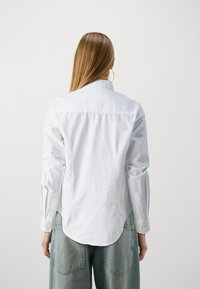 Camicia bianca in cotone con maniche lunghe, polsini abbottonati e orlo curvo. Abbinata a pantaloni di jeans blu chiaro, vista da dietro e capelli lunghi.