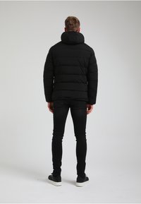 Czarna puchowa kurtka z wysokim kołnierzem i kapturem, prezentująca pikowaną konstrukcję. W zestawieniu z dopasowanymi czarnymi dżinsami oraz ciemnymi sneakerami.