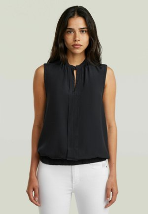 Junge Frau mit langen dunklen Haaren und Sommersprossen trägt eine ärmellose schwarze Bluse und weiße Hose vor einem schlichten Hintergrund.