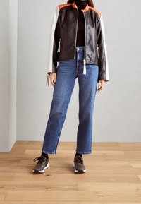 Osoba oblečená v černé a oranžové kožené bundě, černém roláku, modrých džínách a hnědých teniskách stojí na dřevěné podlaze.