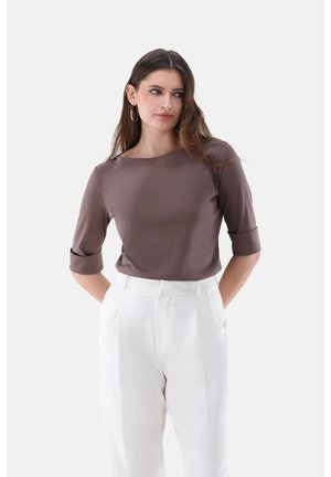 Frau mit langen braunen Haaren, die ein mauvefarbenes Dreiviertelarm-Oberteil und hochtaillierte weiße Hosen trägt, steht mit den Händen in den Taschen.