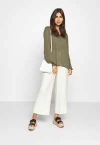 Olive green long-sleeve blouse with a subtle sheen, paired with creamy wide-leg trousers. Black platform sandals and a white crossbody bag.