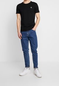 Muž v černém tričku Calvin Klein, modrých džínách s manžetami, s bílými teniskami, stojící s jednou rukou v kapse proti jednoduchému bílému pozadí.