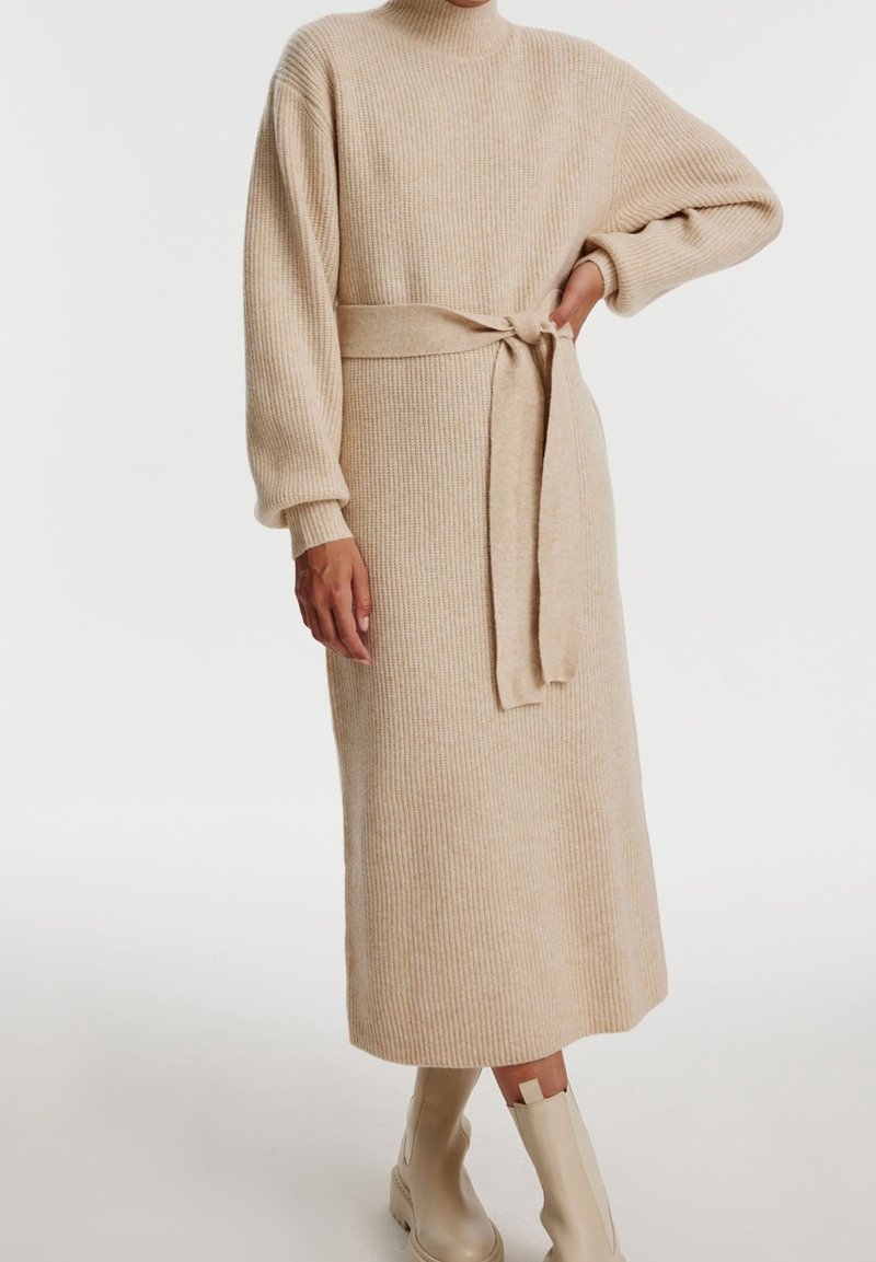 Robe midi en maille de couleur beige clair avec une texture côtelée, un col montant, des manches longues et une ceinture assortie à la taille. Portée avec des bottes de couleur crème.