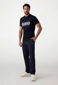 Fekete póló fehér "GUESS" logóval, sötét szabadidőnadrággal és fehér edzőcipővel párosítva. A modell egyik kezét a zsebében tartja, semleges háttér előtt áll.