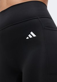 Gros plan sur un legging de sport noir avec un logo Adidas blanc près de la hanche et des coutures visibles sur un tissu lisse.