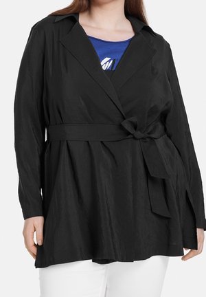 Persoon draagt een zwart jasje met riem over een blauw top en witte broek, met het jasje in een knoop gestrikt ter hoogte van de taille.