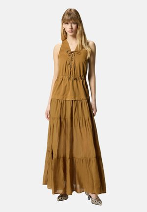 Femme debout, portant un top marron sans manches à lacets avec une taille froncée et une jupe maxi à volants assortie, associée à des talons effet peau de serpent.