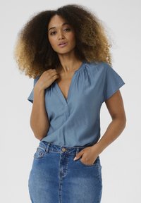 Blouse bleu clair avec un col en V, des manches courtes et un détail froncé au niveau du col. Associée à une jupe en denim taille haute.