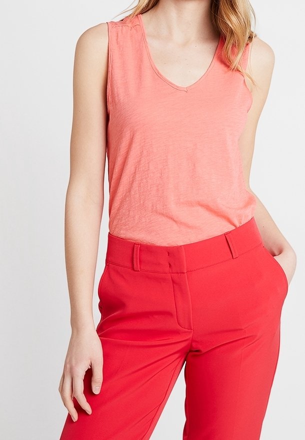 Mujer con una blusa sin mangas de pico en color coral y pantalones rojos de cintura alta, con una mano en el bolsillo y la otra relajada a un lado.