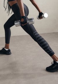 Czarne legginsy sportowe posiadają wyraźne logo "PUMA" z boku, zestawione z dopasowanym czarnym topem na ramiączkach i rękawicami, trzymając srebrne hantle.