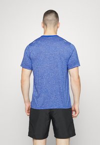 Maglietta atletica blu con maniche corte, tessuto strutturato, scollatura rotonda e dettagli riflettenti, abbinata a pantaloni corti neri. Vista posteriore.