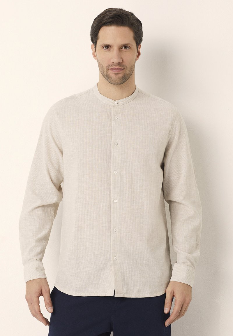 Camicia a maniche lunghe, beige chiaro, con colletto alla cinese, texture sottile e chiusura frontale con bottoni. Indossata sopra pantaloni scuri.