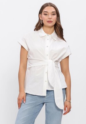 Vrouw die een wit overhemd met korte mouwen en knopen draagt met een strik aan de voorkant en lichtblauwe spijkerbroek, staand tegen een effen witte achtergrond.