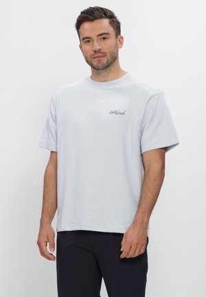 Man met kort bruin haar die een lichtblauw oversized T-shirt en een zwarte broek draagt, staand tegen een witte achtergrond.