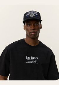Férfi fekete "Les Deux" sapkában és fekete pólóban, fehér felirattal egy sima világos háttér előtt, szemben állva, semleges arckifejezéssel.