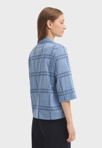 Chemise bleu clair à manches courtes avec un motif en quadrillage et une broderie ajourée. Ourlet arrondi et silhouette simple, fabriquée en coton.