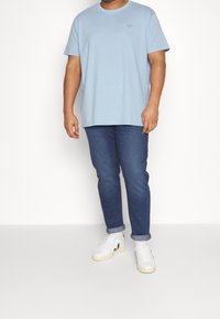 T-shirt in cotone azzurro chiaro con maniche corte, abbinato a jeans blu scuro e sneakers bianche. La t-shirt presenta un branding sottile.