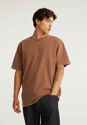 Młody mężczyzna o ciemnych kręconych włosach, ubrany w brązowy t-shirt z krótkim rękawem i ciemne jeansy, patrzący na bok z jedną ręką w kieszeni.