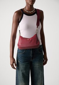 Red leather shoulder bag with a curved shape, featuring a textured surface, gold-tone hardware, and a patterned strap. Casual denim pants.