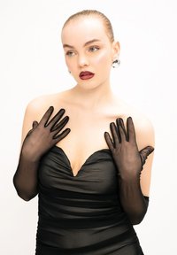 Guanti sheer neri accentuano un vestito aderente nero con profondo scollo a V. La modella ha un aspetto liscio e lucido con trucco neutro.