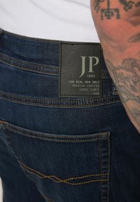 Jeans in denim blu scuro con cuciture ben visibili. Una toppa in pelle nera mostra "JP 1880" e "PER UOMINI VERI SOLO" in lettering argentato.