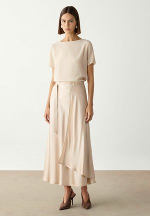 Kvinde med lige brunt hår iført beige kortærmet top, matchende midilang nederdel med knude foran og brune spidse høje hæle, stående på beige gulv.