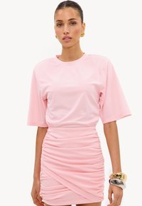 Femme portant une mini-robe rose clair froncée à manches courtes, boucles d’oreilles créoles en or, et bracelets épais en or et argent à son poignet droit.